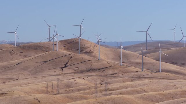 Aerial shot captures wind turbines rotating across dry rolling hills under a clear sky in California, generating clean sustainable electricity across an expansive renewable wind power farm.