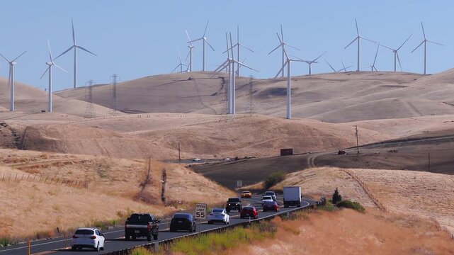 Vehicles travel along a curving two lane highway winding through arid rolling hills, past electric power lines and rotating wind turbines under clear daytime skies in Altamont Pass, California.