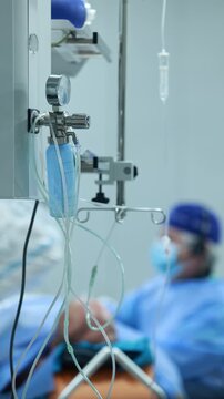 Oxygen flowmeter with humidifier and IV drip in hospital operating room, blurred patient on table and surgeon in background during cardiac procedure