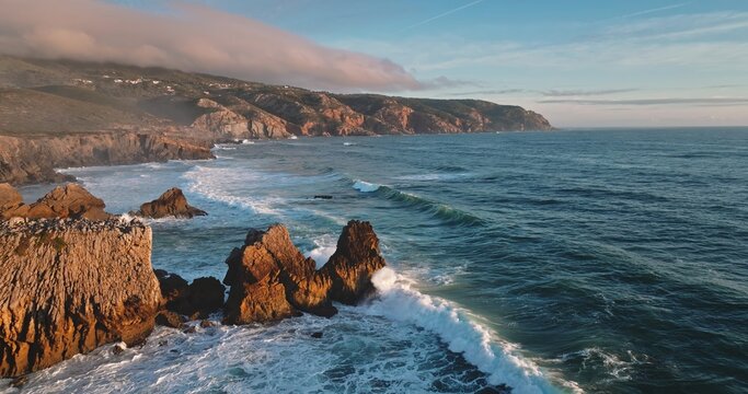 Aerial view of the rugged coastline of Lisbon with dramatic cliffs, crashing waves, and golden hour light hitting the rocky formations in Lisbon, Lisbon, Portugal.