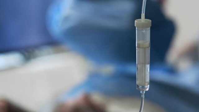 Close up of IV drip chamber with falling drop in hospital operating room, blurred surgical team and surgical light in background, copy space