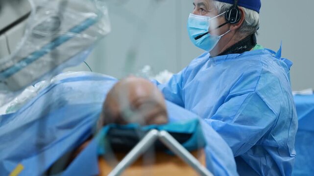 Experienced senior surgeon with headset performing cardiac catheterization in hospital cath lab, patient on operating table and C-arm in foreground