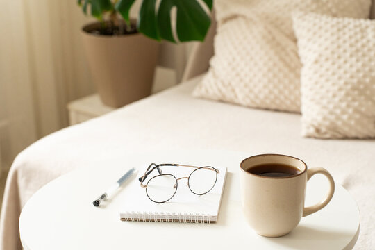 An atmosphere of coziness and measured pace of life, cup of coffee, notebook, pen, and glasses on table in light-filled room. Mindfulness, journaling, me-time, and calm daily routine