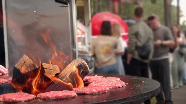 Close up street food chef holds stack burger buns. This medium shot shows gloves clad hands arranging buns for a firepit style griddle. Many raw patties are visible next to a large flame.