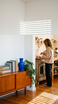 Young woman cooking homemade food on the stove in a cozy kitchen viewed from a stylish sunlit living room