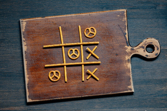 Tic tac toe game made of pretzel snacks, closeup. Creative tic tac toe game board made with salty pretzel sticks and twists on rustic wooden cutting board over dark background
