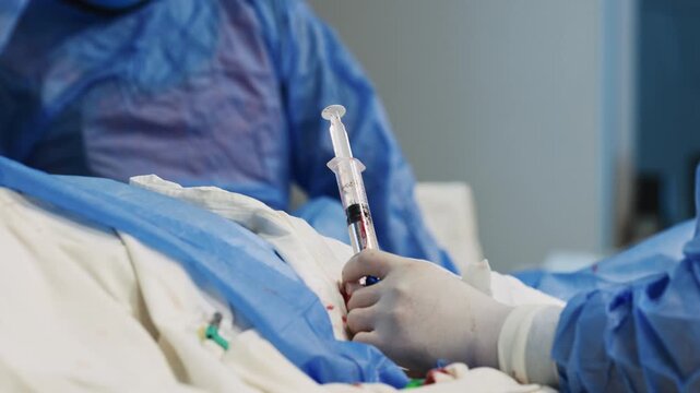 Interventional cardiology procedure, doctor working with catheter and syringe over sterile draped patient while colleague follows angiography screen