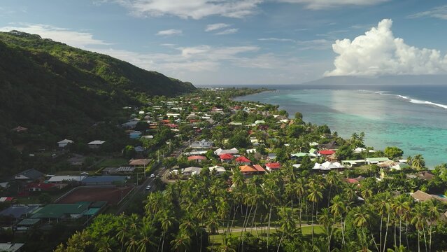Aerial view of the coastal town of Moorea-Maiao nestled between lush green mountains and a turquoise lagoon, with palm trees and houses in Moorea-Maiao, Windward Islands, French Polynesia.