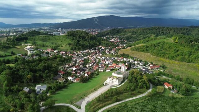 Aerial drone view of Blessed Anton Martin Slomsek Church in Kosaki, Maribor, surrounded by homes and vineyards, with Maribor and the forested Pohorje range in the background.

