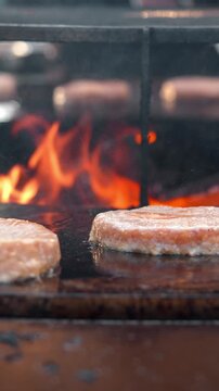 Vertical frame shows appetizing meat burger patties grilling on hot griddle surface against bright open flame background. Juicy minced meat cooks during outdoor culinary festival.