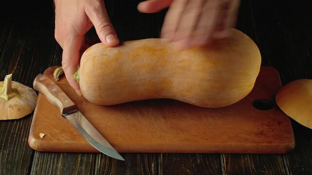 Hands rotate whole butternut squash on wooden cutting board beside kitchen knife and sliced produce in rustic kitchen food preparation setting