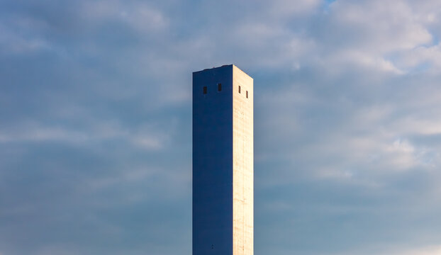 Aerial view of a tall concrete industrial chimney rising above a manufacturing complex against dramatic clouds and morning sky in Brescia, Lombardy, Italy.