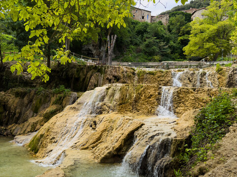peaceful waterfall in rural landscape with stone steps and green trees, serene nature scene with flowing water and stone buildings, side view of tranquil river with stone steps and greenery.