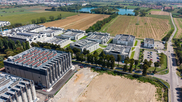 Aerial view of a large data center campus featuring multiple industrial buildings with rooftop cooling systems, parking areas, and surrounding green fields in Milan, Lombardy, Italy.