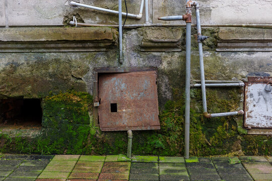 Neglected utility facade with rusted panels, exposed pipes, moss, damp masonry and corrosion. Aging building infrastructure requiring maintenance