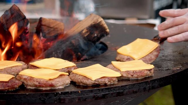 Cook flips juicy burgers with bright yellow cheese on flat pan. Busy carnival food stall relies on massive central wood burning pit to prepare thick fresh hot sandwiches for crowds.