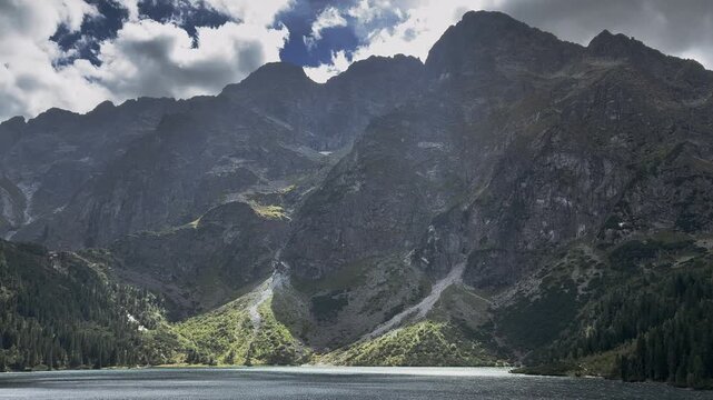 Majestic mountain lake surrounded by rugged rocky peaks and green forest under dynamic cloudy sky. Beautiful alpine nature landscape. Morskie oko 