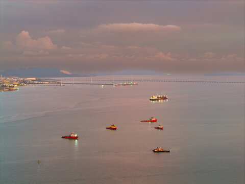 Aerial view of cargo ships and tugboats navigating the calm waters of the Penang Strait with the Penang Bridge in the distance under a soft sunset sky in George Town, Penang, Malaysia.