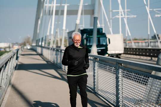 Active elderly male in sportswear running across a bridge pathway for cardiovascular health and morning workout routine.