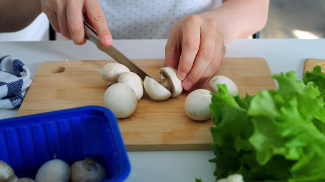 Woman slices fresh champignon mushrooms using knife on wooden cutting board, preparing healthy ingredients near green lettuce leaves with focused calm precision during wholesome culinary routine.
