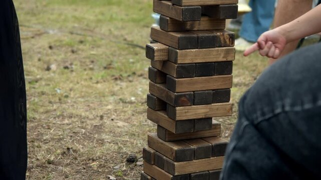 Hand pulls wooden block from giant tower game. People enjoy outdoor entertainment at summer street food fair.