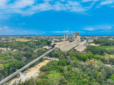 Aerial view of a large industrial cement factory complex surrounded by dense green forests and trees under a bright blue sky in Cilacap, Central Java, Indonesia.