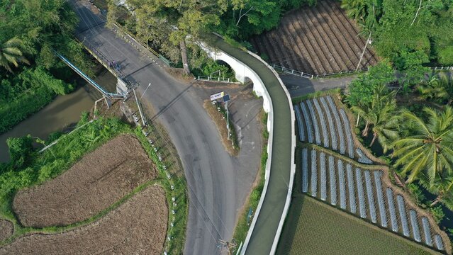 Aerial view of an irrigation aqueduct bridge spanning over a road surrounded by agricultural fields and palm trees in Tempel, Special Region of Yogyakarta, Indonesia.