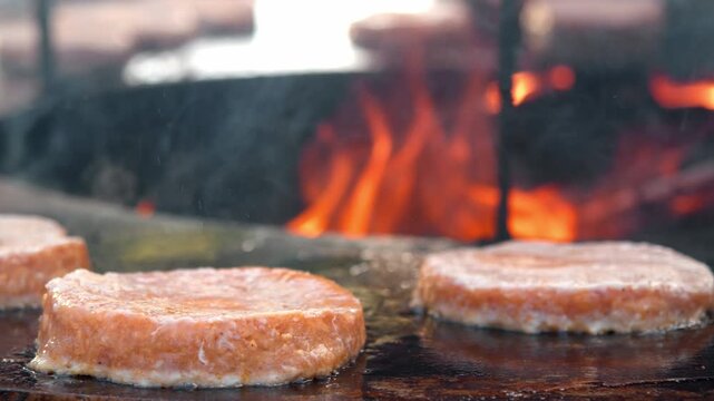 Close up shows raw meat burger patties cooking on hot flat grill. Bright open flame burns in background while juicy pork roasts at street food festival.