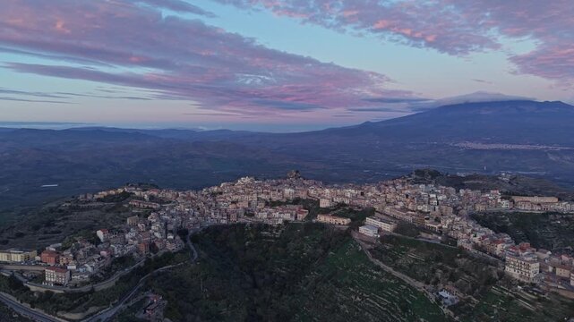 Aerial view of the historic town of Centuripe perched on a hilltop with Mount Etna in the background under a soft sunset sky in Centuripe, Sicily, Italy.