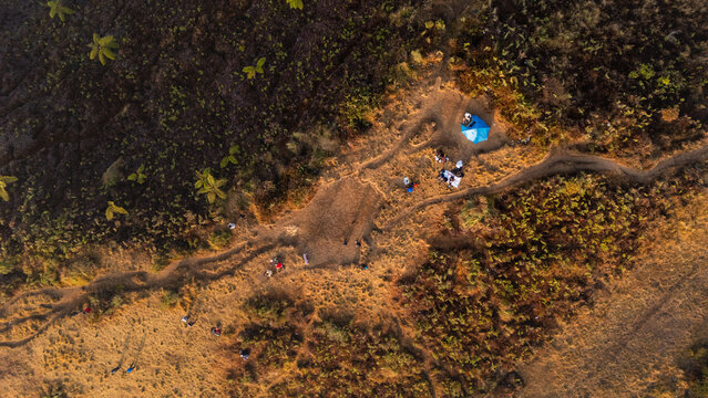 Aerial view of the Sunrise Camp Gunung Prau area with a blue tent and hikers on a mountain trail surrounded by dry grassy terrain in Kecamatan Kejajar, Jawa Tengah, Indonesia.