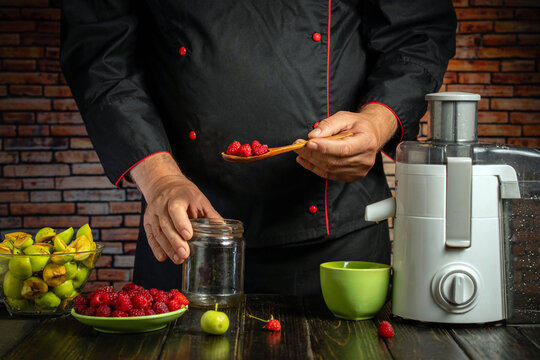Cook holds spoonful of strawberries beside blender and open jar. Black apron frames hands preparing fruit, with chopped apples and berries arranged on dark kitchen counter for blending into drink.