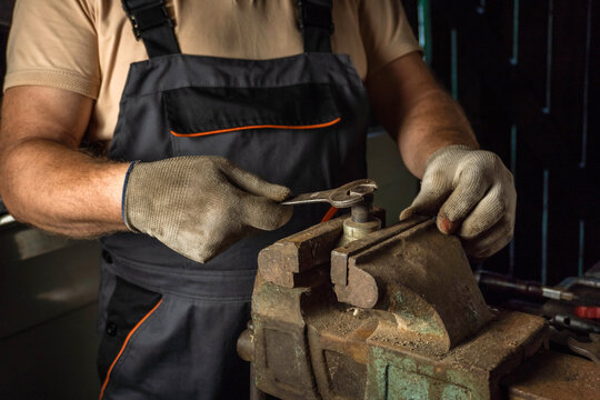 In a rustic workshop filled with tools, a skilled worker in gloves tightens a metal piece with a wrench. The focus is on precision and craftsmanship in the trade