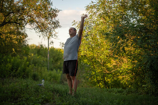 Man holds overhead stretch with elastic band on grassy woodland trail. Fitness exercise looks steady and concentrated, with bare feet, gray shirt and quiet natural setting around him.