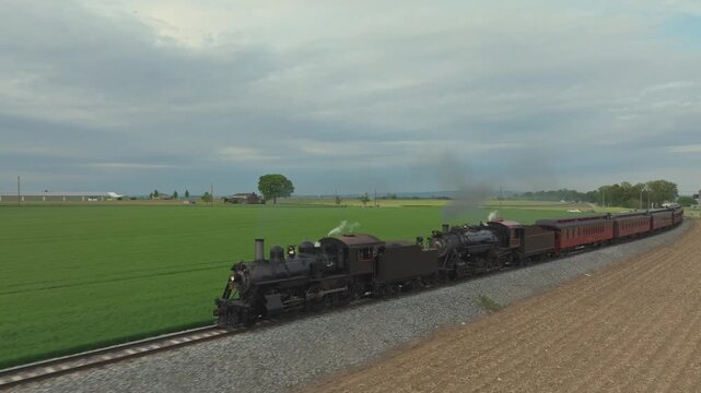 A steam train moves along railway tracks through a green field. It passes by trees and open land with a gentle smoke plume. The sky is cloudy, creating a muted light.