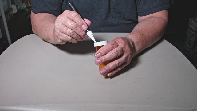 A person uses a marker to write on a container while sitting at a table. The hands move clearly as the writing progresses. The setting shows a simple workspace with a neutral background.