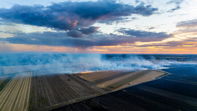 Aerial view of a field fire burning in agricultural land with smoke rising against a dramatic sunset sky in Sremska Mitrovica, Vojvodina, Serbia.