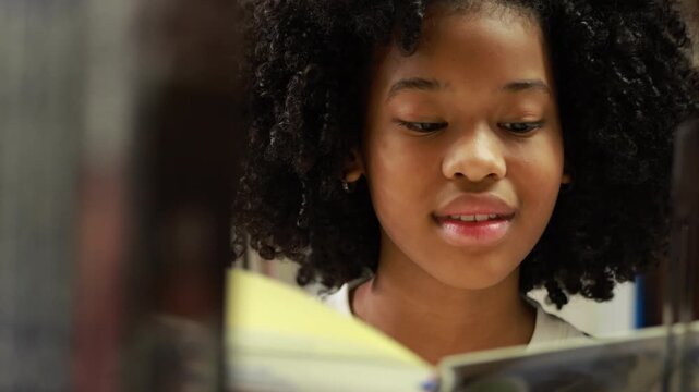 Young girl smiles warmly behind bookshelf. Curly hair frames her cheerful expression. She sits in quiet, cozy library space. Image evokes joy, wooden shelves hold many books. Education, Back to school