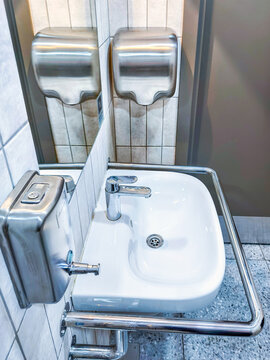 Accessible sink with grab bars in public restroom. Stainless steel soap dispenser and paper towel dispenser. Handwashing and hygiene. Barrier-free design and disability access