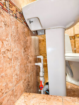 Pedestal sink with plumbing pipes and water valve under washbasin in tiled bathroom. Toilet in background. Bathroom renovation and repair work. Plumbing installation concept