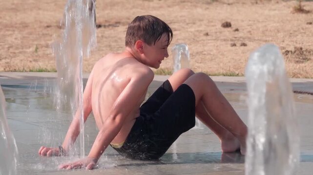 A joyful boy cools down and enjoys playing under fresh splashing jets of a public ground fountain on a bright hot sunny afternoon.