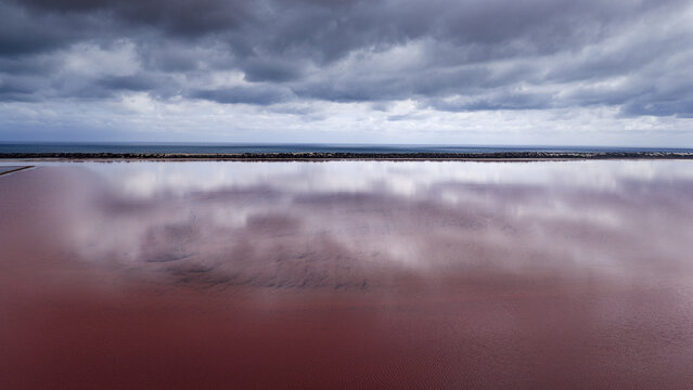 Aerial view of a vibrant pink salt lake with dramatic dark clouds reflecting on the water surface and a distant coastline in Australia.