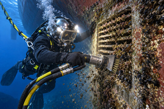 A commercial diver uses a powered brush to remove marine growth from a ship hull grille, surrounded by bubbles and floating debris in blue water.
