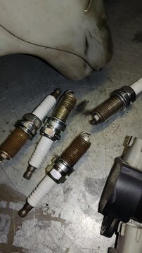 Close-Up View of Worn Spark Plugs with Corroded Electrodes on Workshop Floor Under Bright Workshop Lighting