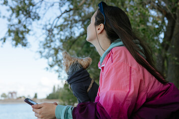 Outdoor lifestyle scene of a white woman in a pink jacket holding a smartphone beside a Yorkshire Terrier near the water, surrounded by green trees, bright daylight, and colorful clothing outside. © vita