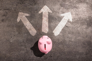 Fototapeta premium Pink piggy bank in front of arrows pointing in different directions on grey stone surface, top view.