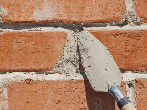 A close up view of a metal trowel applying grey mortar between weathered red clay bricks during a masonry wall construction or repair process