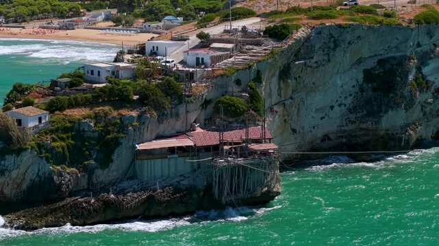 Aerial reveal of Trabucco da Elia on cliff promontory Peschici Gargano Puglia 70mm telephoto drone pull back shot showing traditional wooden fishing structure over Adriatic Sea with ambient wave sound