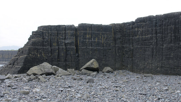 A tall, dark, vertical cliff face with distinct horizontal and vertical rock layers, rising above a rocky beach with scattered stones and pebbles
