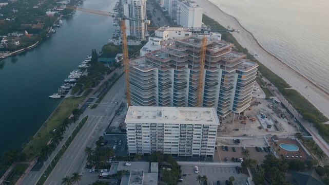 Aerial view of a large modern building under construction with tall cranes, located along a canal with docked boats and the sandy coastline in Miami Beach, Florida, United States.