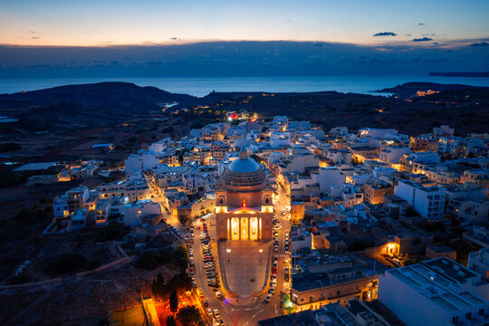 Aerial view of the Parish Church of the Assumption of the Blessed Virgin Mary into Heaven illuminated at dusk, surrounded by the town of L-Imgarr, Regjun Tramuntana, Malta.